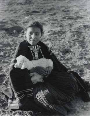 A black-and-white photograph of an Indigenous girl sitting on the ground while holding a lamb.