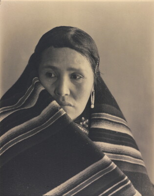A black-and-white portrait photograph of an Indigenous woman with a blanket wrapped around her neck and shoulders.