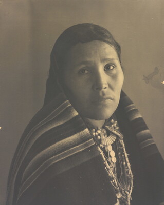 A black-and-white portrait photograph of an Indigenous woman with a blanket wrapped around her neck and shoulders.