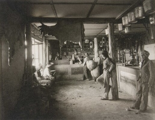 A black-and-white photograph of Indigenous people in a shop with a variety of products such as animal hides, tin pails, and lanterns hanging from the ceiling.