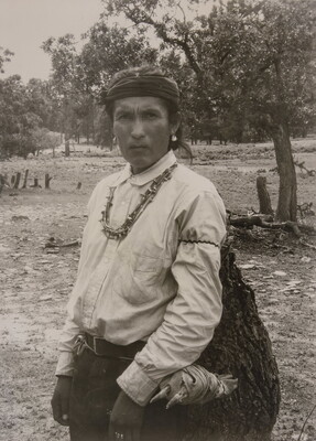 A sepia-toned photograph of an Indigenous man carrying a large bag.