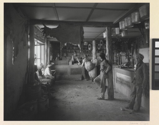 A black-and-white photograph of Indigenous people in a shop with a variety of products such as animal hides, tin pails, and lanterns hanging from the ceiling.