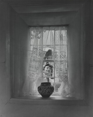 A black-and-white photograph of a sheer curtained window with a Native American-style pot on the sill as a White woman cleans the window from outside.