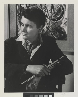 A black-and-white portrait photograph of a seated White woman holding paintbrushes in front of an abstract painting.