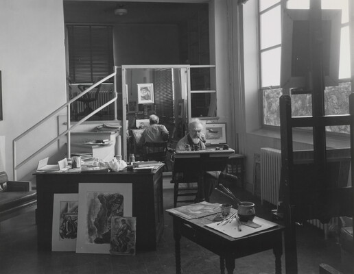 A black-and-white photograph of an older White man smoking a pipe seated at a desk working; he is surrounded by art supplies and artwork on tables, and a mirror behind him that shows him from the back.