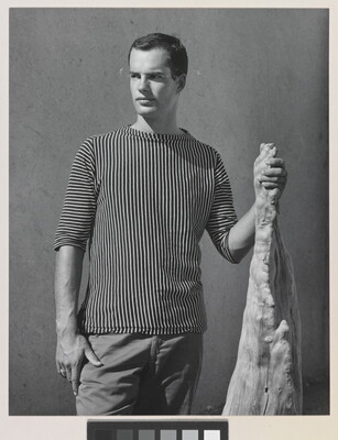 A black-and-white photograph of a White man with wearing a shirt with vertical stripes and holding a wooden object.