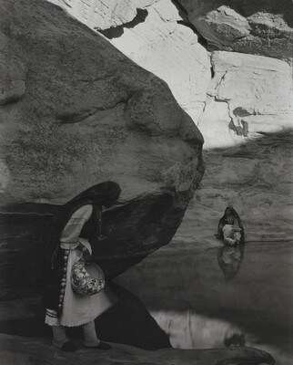 A black-and-white photograph of a woman with a long headscarf standing at the edge of a water hole surrounded by boulders and rock walls.