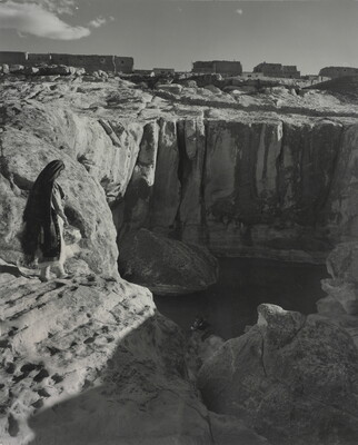 A black-and-white photograph of a woman with a long headscarf on a cliff, overlooking a water hole at the bottom.