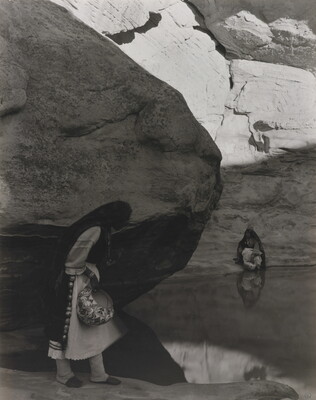 A black-and-white photograph of a woman with a long headscarf standing at the edge of a water hole surrounded by boulders and rock walls.