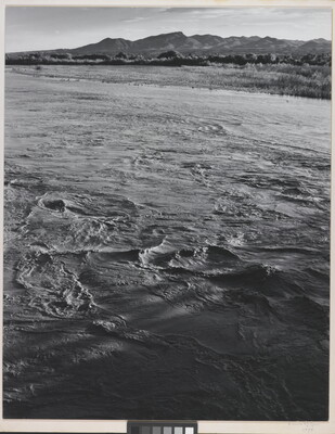 A black-and-white photograph of a choppy river and then grassy plains and mountains beyond the shore.