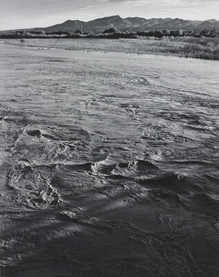 A black-and-white photograph of a choppy river and then grassy plains and mountains beyond the shore.