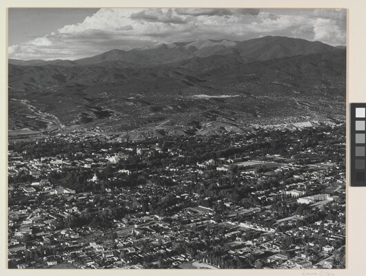 A black-and-white aerial photograph of a town in a mountain valley beneath puffy white clouds.
