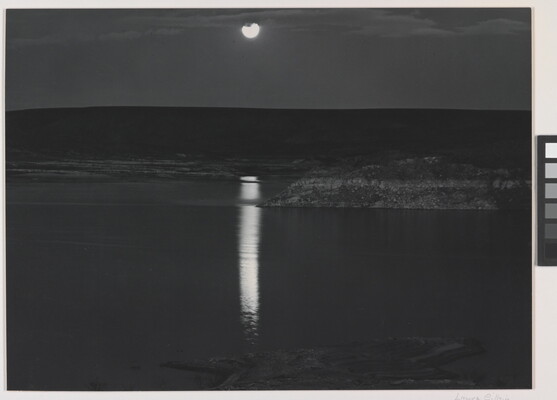 A black-and-white photograph of a night sky and full moon reflected across calm water.