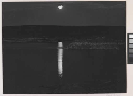 A black-and-white photograph of a night sky and full moon reflected across calm water.