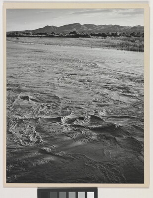 A black-and-white photograph of a choppy river and then grassy plains and mountains beyond the shore.
