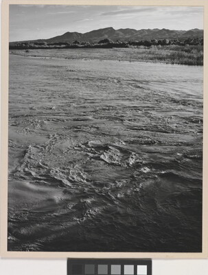 A black-and-white photograph of a choppy river and then grassy plains and mountains beyond the shore.
