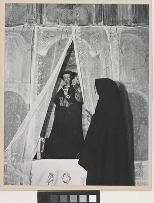 A black-and-white photograph of a woman wearing a black shawl over her head facing a statue of a saint holding a large key.