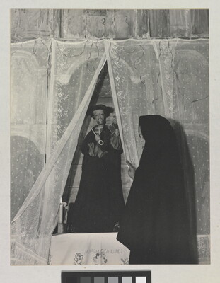 A black-and-white photograph of a woman wearing a black shawl over her head facing a statue of a saint holding a large key.