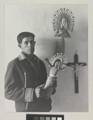 A black-and-white photograph of a medium-skinned man in a winter jacket holding an elaborately carved wooden figure.