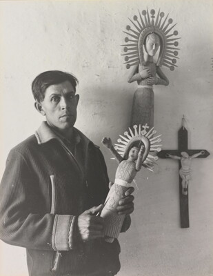 A black-and-white photograph of a medium-skinned man in a winter jacket holding an elaborately carved wooden figure.