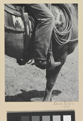 A black-and-white photograph of a person's leg in a cowboy boot, including spurs, sitting astride a horse, viewing only the horse's front quarter.