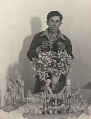 A black-and-white photograph of a medium-skinned man standing behind a wood sculpture of trees and human figures.