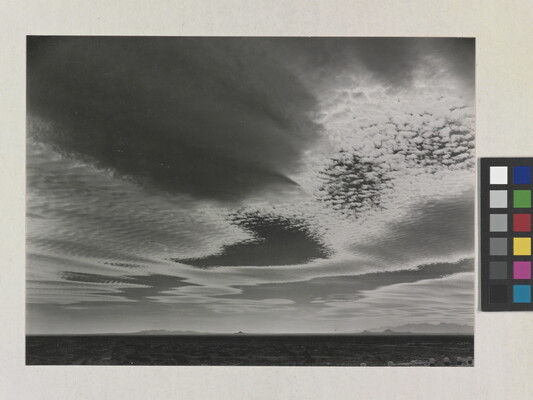A black-and-white photograph of an expansive desert plain under rippled clouds with mountains in the far distance.