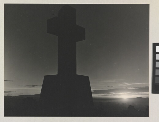 A black-and-white photograph of a stone cross in silhouette and the sun peeking out from the horizon.