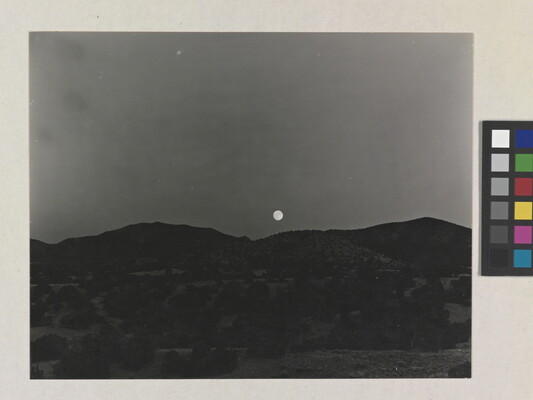 A black-and-white photograph of the moon rising above a mountain landscape.