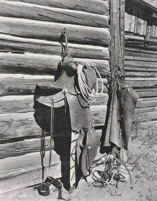 A black-and-white photograph of a leather saddle, chaps, rope, and other horse gear hung on the side of a log building.