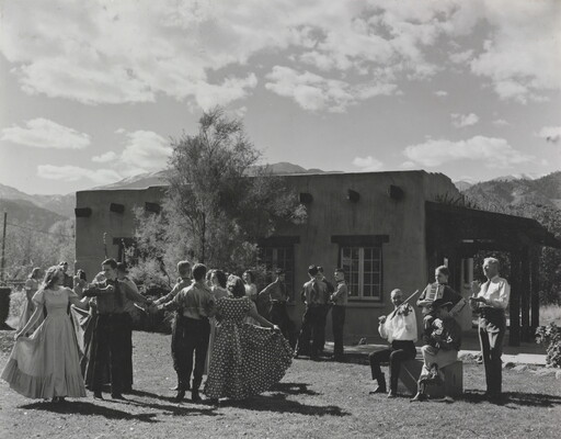 A black-and-white photograph of groups of people dancing outside as a band plays in front of a low adobe building.