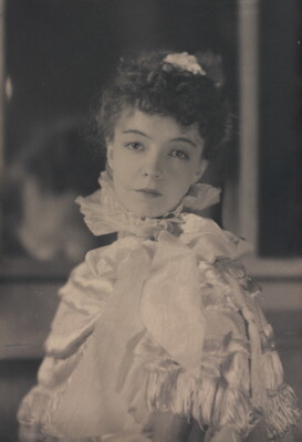 A black-and-white portrait photograph of a White woman with curly hair pulled on top of her head, wearing a top with a ruffled collar.