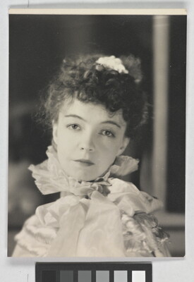 A black-and-white portrait photograph of a White woman with curly hair pulled on top of her head, wearing a top with a ruffled collar.