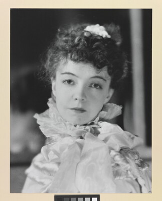 A black-and-white portrait photograph of a White woman with curly hair pulled on top of her head, wearing a top with a ruffled collar.