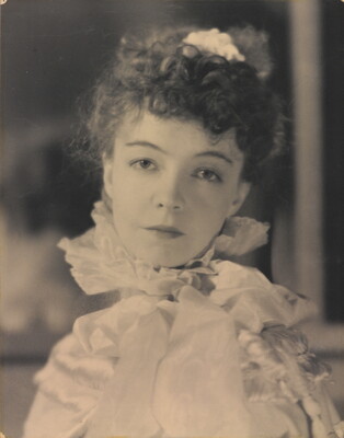 A black-and-white portrait photograph of a White woman with curly hair pulled on top of her head, wearing a top with a ruffled collar.