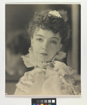 A black-and-white portrait photograph of a White woman with curly hair pulled on top of her head, wearing a top with a ruffled collar.