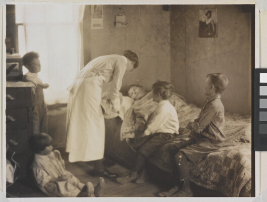 A black-and-white photograph of a nurse bending over a person in a bed, two children sitting on the bed, and two children behind the nurse.