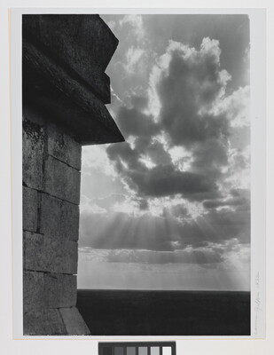 A black-and-white photograph of the sun's rays bursting from clouds and a stark brick architectural feature in the left foreground.