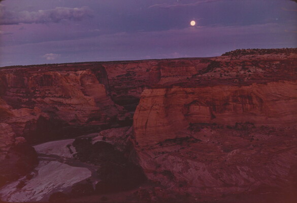 A color photograph of a full moon shining in a purple and blue sky over a steep, red, rocky canyon.
