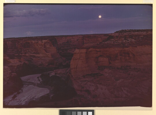 A color photograph of a full moon shining in a purple and blue sky over a steep, red, rocky canyon.