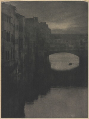 A sepia-toned photograph of a wall of tall buildings lining a river and an enclosed arched bridge stretching over the water.