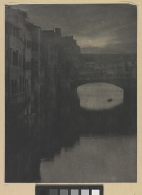 A sepia-toned photograph of a wall of tall buildings lining a river and an enclosed arched bridge stretching over the water.