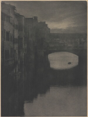 A sepia-toned photograph of a wall of tall buildings lining a river and an enclosed arched bridge stretching over the water.