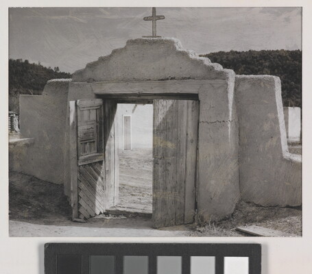 A black-and-white photograph of an adobe wall with a doorway that leads to a courtyard; a cross on the wall above the wooden doors.