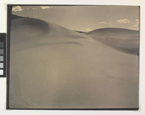 A sepia-toned photograph of a rippling sand dune rising up into a sky dotted with a few small clouds.