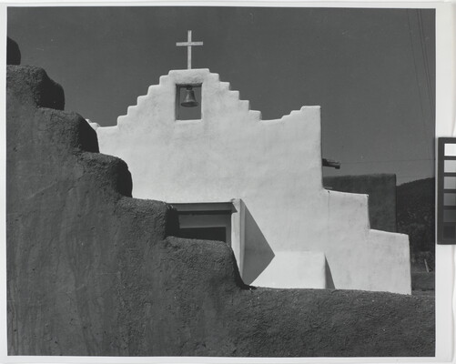 A black-and-white photograph of a terraced adobe bell tower with a cross on top, partially hidden by a terraced adobe wall.