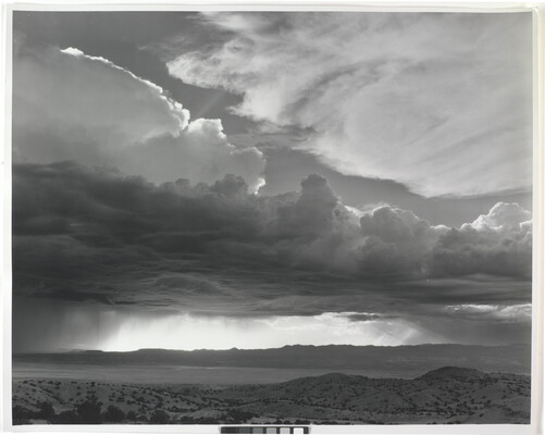 A black-and-white photograph of torrential rain and dark, heavy storm clouds passing into a brightly lit plain.