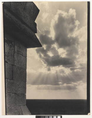 A sepia-toned photograph of the sun's rays bursting from clouds and a stark brick architectural feature in the left foreground.