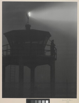 A black-and-white photograph of a person standing in silhouette atop a tower with a bright spotlight shining from the top.