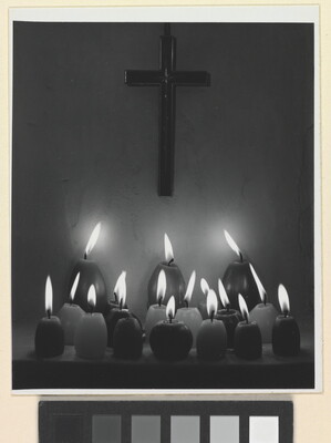A black-and-white photograph of three rows of lit votive candles in front of a cross hanging on a wall.